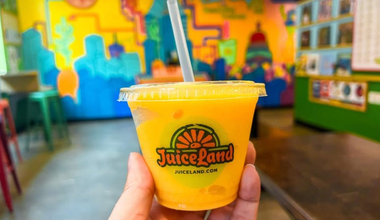 Hand holding a small clear plastic cup of bright orange mango smoothie with lid and straw, blurred colorful mural and stools in a vibrant indoor juice bar.