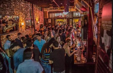 Crowded brick-walled downtown bar at night with warm lighting, exposed beams, a busy wooden bar stacked with drinks and patrons, and a packed standing crowd for lively city nightlife.