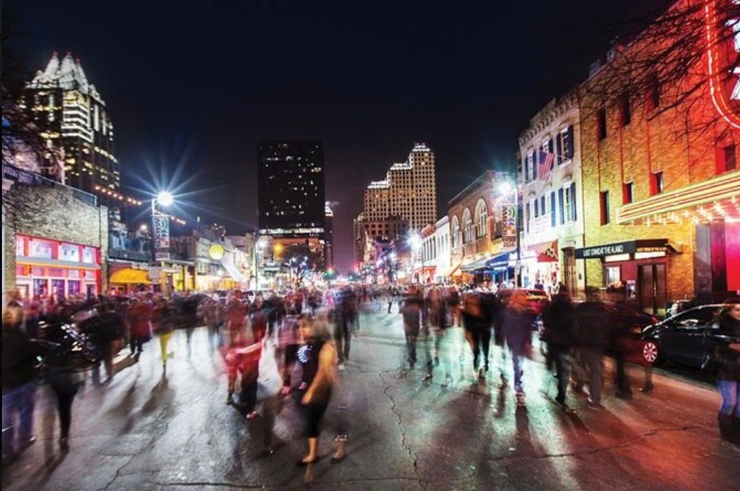 Bustling downtown nightlife street at night with blurred pedestrians, colorful neon signs and theater marquees, wet pavement reflecting city lights and skyline towers in the distance.