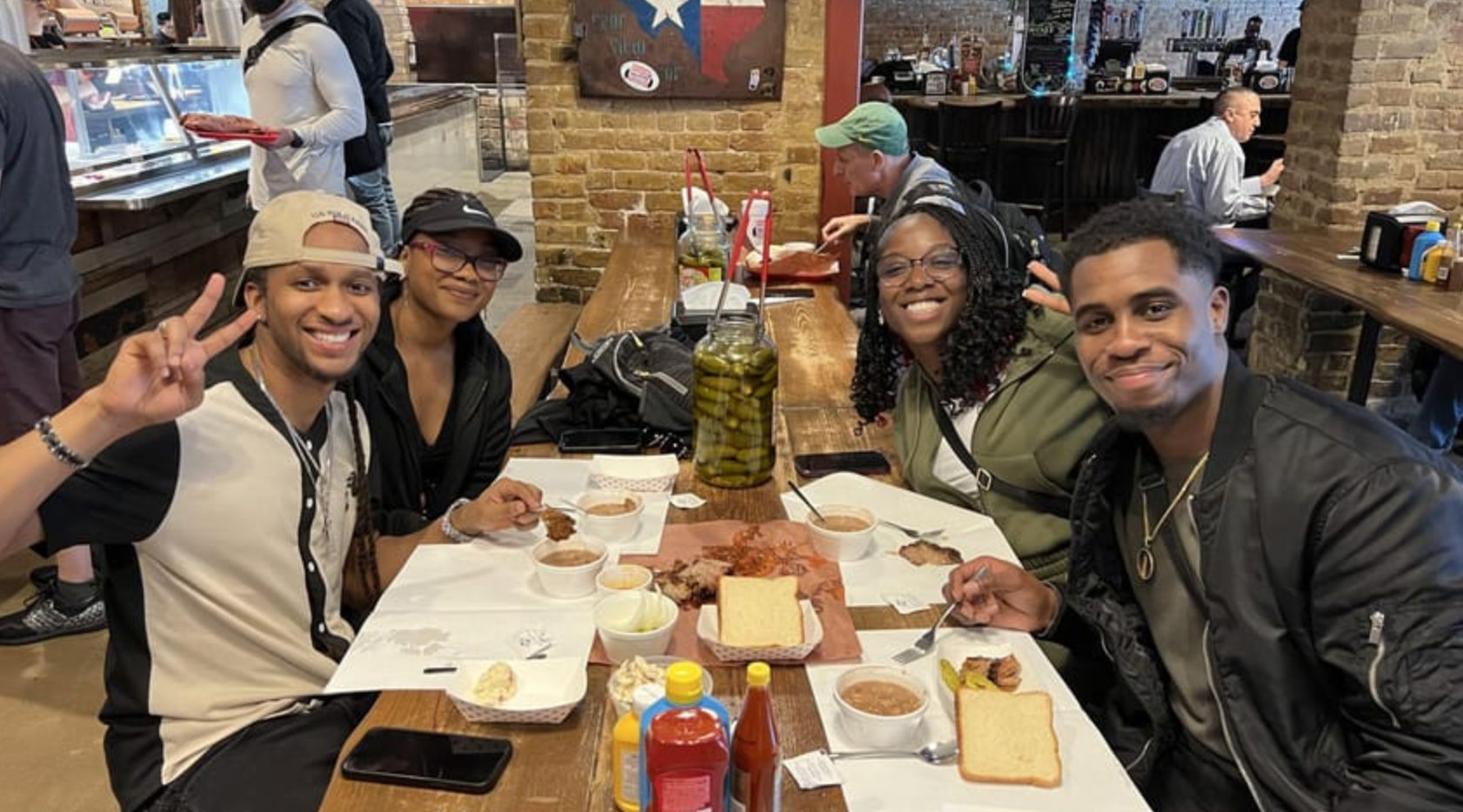 Four friends smiling at a wooden communal table in a brick-lined BBQ joint, sharing plates of smoked meat, slices of white bread, sides, condiments and a large jar of pickles.