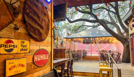 Rustic wooden outdoor bar patio under a large oak tree with string lights, canopy-covered picnic tables, vintage soda signs and mounted antlers