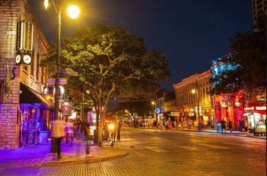 Vibrant downtown entertainment district at night — colorful bars and restaurants along a tree-lined cobblestone street with pedestrians, neon signage, and string lights creating a lively nightlife atmosphere
