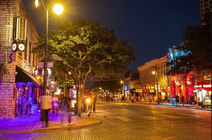 Vibrant downtown entertainment district at night — colorful bars and restaurants along a tree-lined cobblestone street with pedestrians, neon signage, and string lights creating a lively nightlife atmosphere