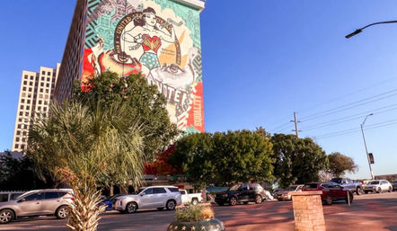 Street-level view of a colorful mural of a stylized female superhero with patriotic motifs on a tall downtown building, framed by palm trees, parked cars, and a clear blue sky.