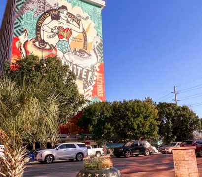 Street-level view of a colorful mural of a stylized female superhero with patriotic motifs on a tall downtown building, framed by palm trees, parked cars, and a clear blue sky.