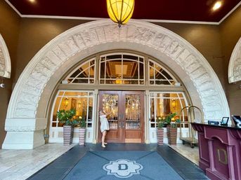 Grand arched stone entrance to a historic hotel with ornate wooden-and-glass double doors, potted plants, bellman's luggage cart, hanging lantern, and a person playfully leaning on the door.