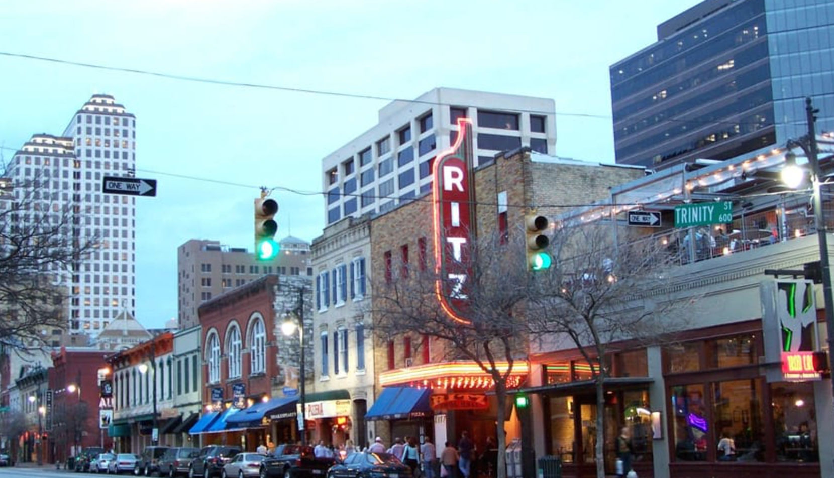 Evening downtown street scene in an urban entertainment district with historic brick storefronts, a vertical neon theater marquee, bustling sidewalk cafes, green traffic lights and a high-rise skyline