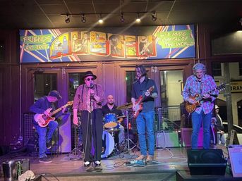 Five-piece live band performing on an intimate indoor stage — vocalist in a hat and sunglasses with drummer and three guitarists under a colorful mural and warm stage lights at a cozy music venue.
