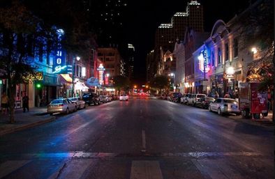 Downtown city nightlife scene at night — neon bar and restaurant signs, parked cars lining both sides, wet reflective street leading to illuminated skyline.