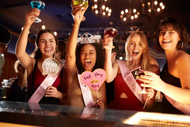Bachelorette party at a bar: four friends in sashes and a tiara cheering with colorful cocktails and photo props saying "She said yes" and "Last fling before the ring".