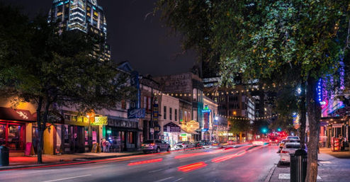 Vibrant downtown nightlife street at night with neon storefronts and historic facades, string lights overhead, red car light trails, parked cars and a lit high‑rise skyline in the background.