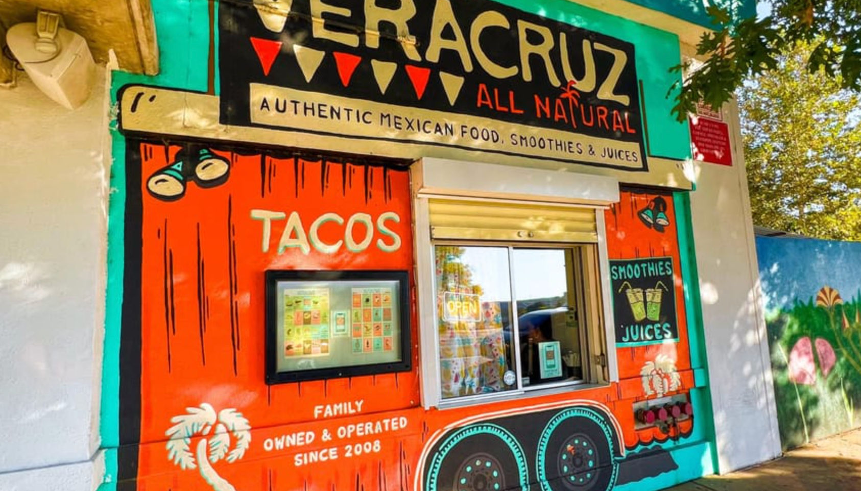 Bright, colorful walk-up taco window with orange-green mural advertising tacos, smoothies and juices — family-run outdoor Mexican takeout.