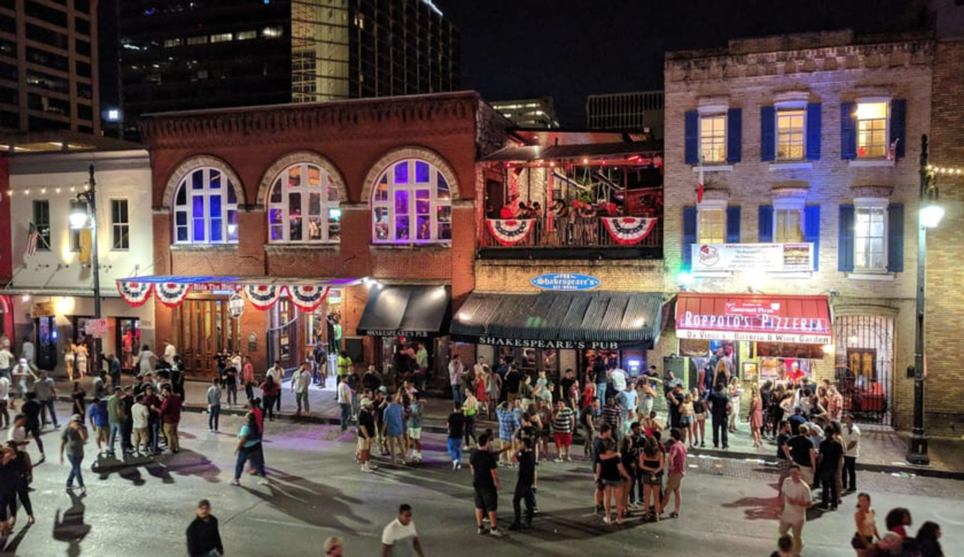 Downtown nightlife scene at night with crowds on the street outside historic brick bars and restaurants, balcony seating, patriotic bunting and colorful window lights.