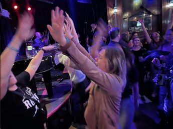 Energetic indoor live-music crowd raising hands toward the stage, drummer visible under blue-purple lighting as people dance and hold drinks in a lively nightlife scene.
