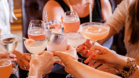 Brunch cocktail toast — friends clinking peach-pink and pale citrus cocktails in coupe and wine glasses, garnishes and manicured hands celebrating.