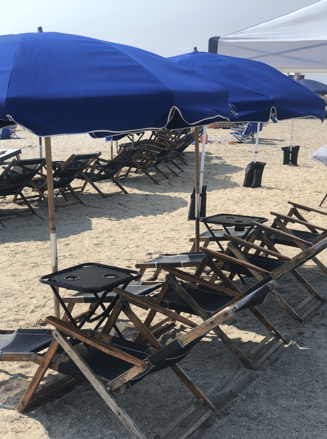 Row of blue beach umbrellas shading wooden folding lounge chairs and small tables on a sandy shore — sunny seaside setup ready for relaxation.
