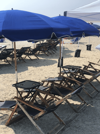 Row of blue beach umbrellas shading wooden folding lounge chairs and small tables on a sandy shore — sunny seaside setup ready for relaxation.