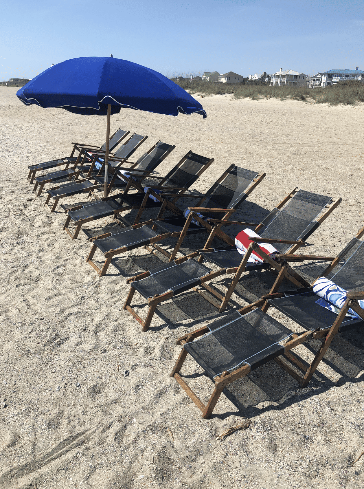 Sunny beachfront scene with a row of wooden lounge chairs and rolled towels under a large blue umbrella on a sandy coastal beach, dunes and beach houses visible in the distance