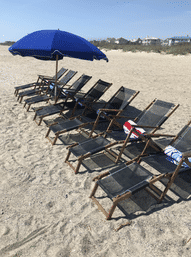 Sunny beachfront scene with a row of wooden lounge chairs and rolled towels under a large blue umbrella on a sandy coastal beach, dunes and beach houses visible in the distance