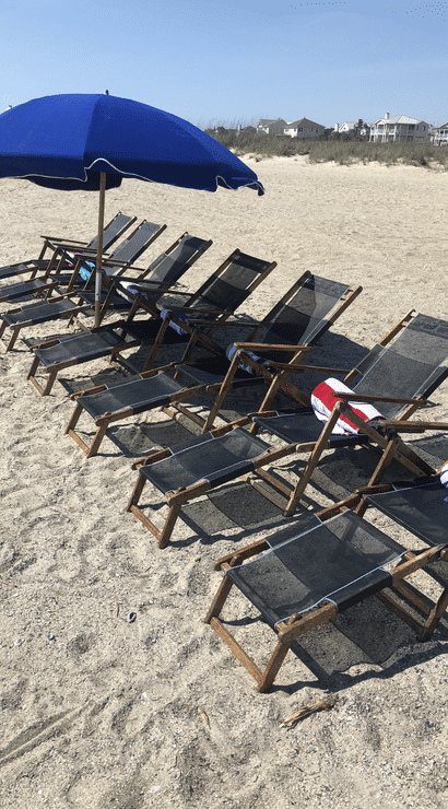 Sunny beachfront scene with a row of wooden lounge chairs and rolled towels under a large blue umbrella on a sandy coastal beach, dunes and beach houses visible in the distance
