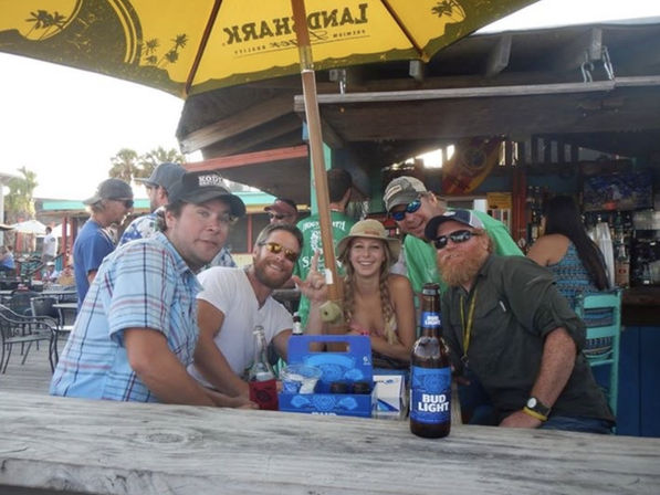 Group of five friends laughing at a sunny beachside tiki bar on a wooden deck under a yellow umbrella, with drinks and a bottled beer on the table.