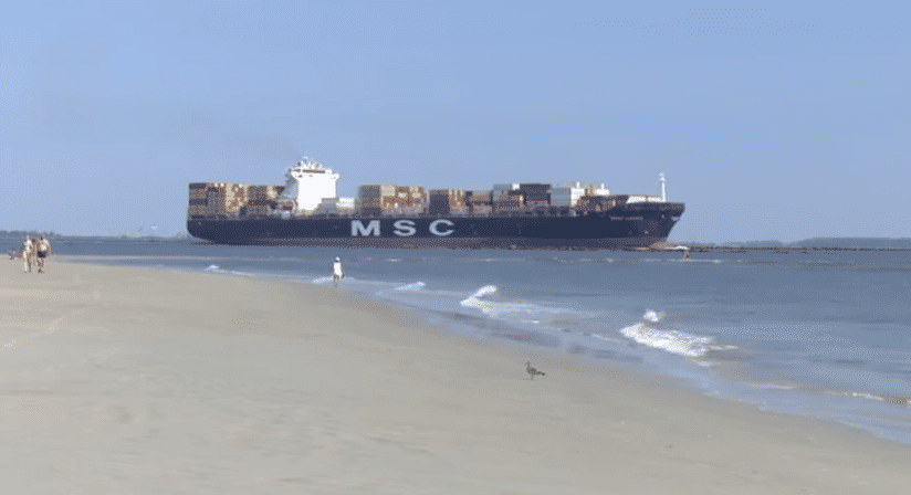 People strolling on a sandy beach shoreline with gentle waves as a large cargo container ship passes offshore under a clear blue sky.
