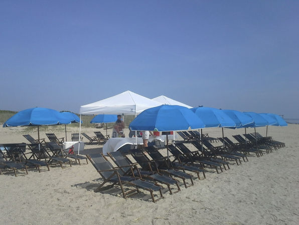 Sandy beach with rows of bright blue umbrellas and wooden lounge chairs lined up near a white canopy beneath a clear blue sky