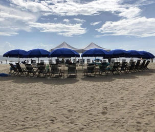 Row of blue beach umbrellas and lounge chairs on sunlit sand, white canopy tents and beachgoers by the ocean under a partly cloudy sky.