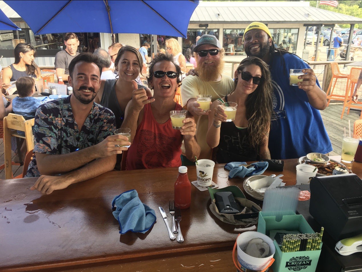 Friends toasting with cocktails at a sunny dockside outdoor bar, seated at a wooden counter under blue umbrellas with drinks, condiments and plates on the table.