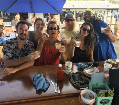 Friends toasting with cocktails at a sunny dockside outdoor bar, seated at a wooden counter under blue umbrellas with drinks, condiments and plates on the table.