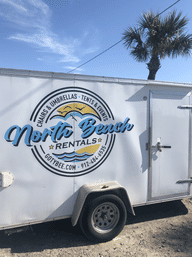 White beach rental trailer with colorful circular logo parked on a sandy lot beneath a palm tree and clear blue sky