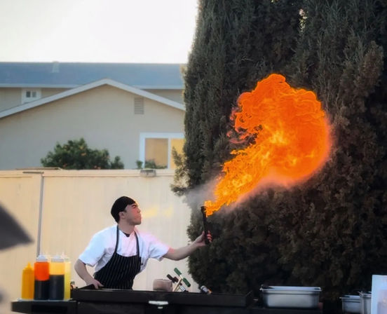 Outdoor chef in white shirt and striped apron using a handheld torch to create a dramatic orange flame plume over a grill station with squeeze bottles nearby.