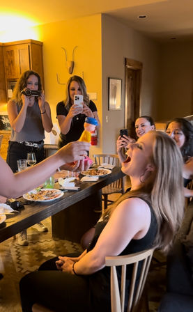 Group of women laughing in a cozy rustic dining room as one seated woman playfully leans back with mouth open while another holds a colorful novelty toy toward her and friends record the moment on phones and a camcorder.