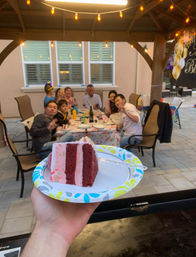 Slice of red velvet cake on a paper plate held in the foreground at a backyard gazebo birthday party, family seated at a decorated table under string lights on a paved patio.