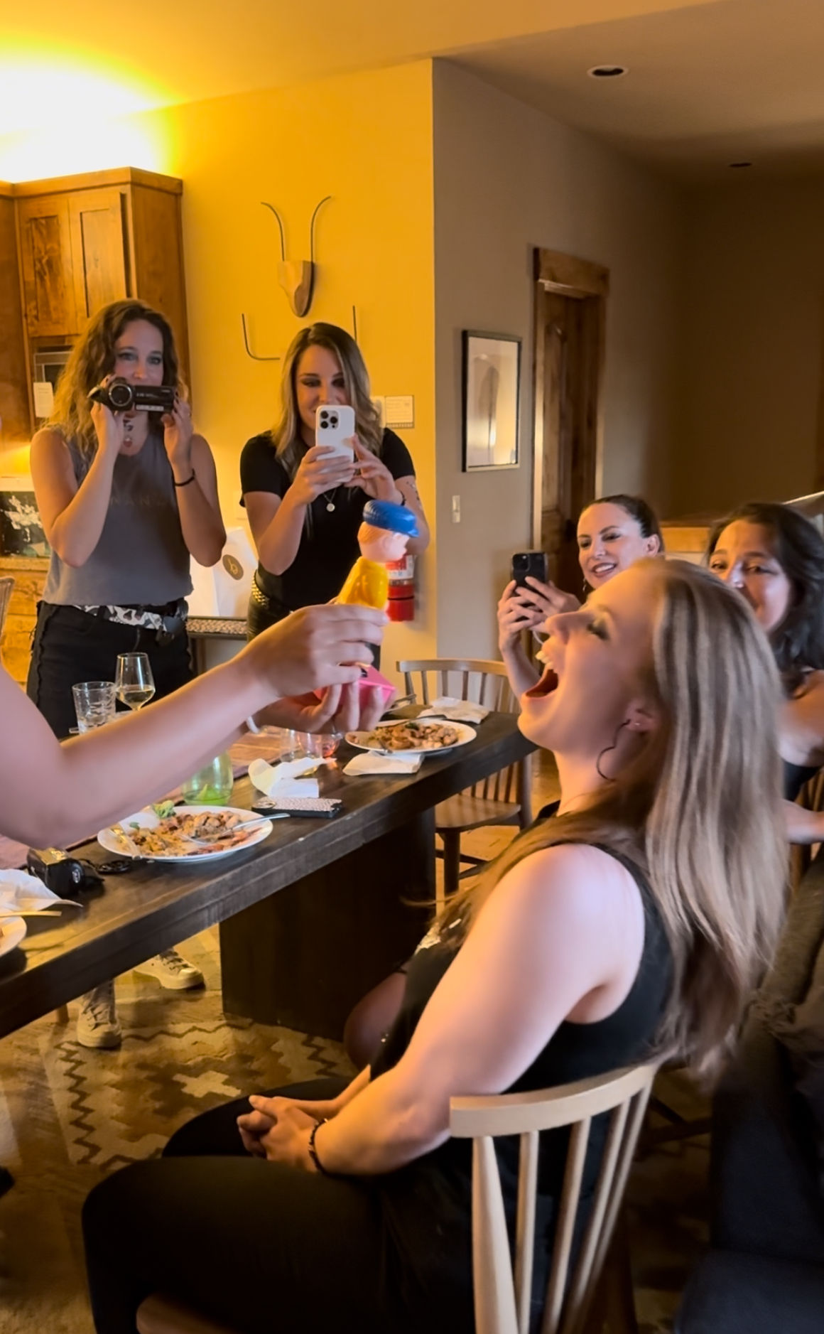 Friends at a cozy cabin-style dining table laughing and recording an indoor group dinner party as someone playfully holds a colorful toy toward a woman who leans back with her mouth open.