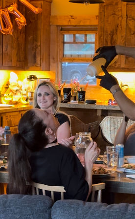 Casual dinner in a cozy wood‑paneled kitchen: friends at a dining table as one person leans back to catch a stream from a yellow squeeze bottle held by gloved hands, with plates, drinks and warm lighting