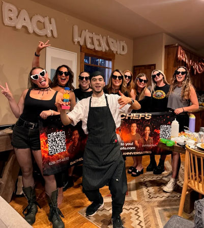 Chef in a striped apron holding a toy, posing with a laughing group of women in heart-shaped sunglasses at a lively bachelorette weekend in a cozy wood-accented home kitchen.