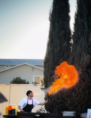 Chef in striped apron producing a dramatic orange flame from a handheld torch over an outdoor grill in a backyard setting with tall cypress trees