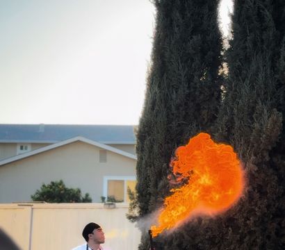 Chef in striped apron producing a dramatic orange flame from a handheld torch over an outdoor grill in a backyard setting with tall cypress trees