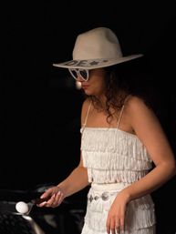 Person in a white fringed dress and wide-brim hat with star decals, wearing rhinestone heart-shaped sunglasses and balancing a white ball on a spatula against a dark background.