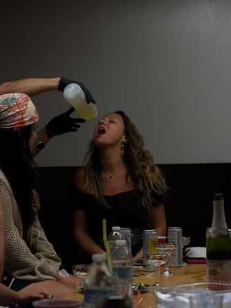Indoor party scene: woman tilting her head back with mouth open as a gloved hand pours a yellow drink from a squeeze bottle, seated at a table scattered with water bottles, cans, champagne and glassware.