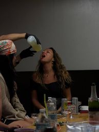 Indoor party scene: a woman leans back with her mouth open as a gloved friend pours a yellow drink from a squeeze bottle; crowded table in foreground with water bottles, cans and champagne glasses.