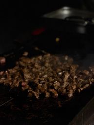 Close-up of sizzling diced steak bites browning on a hot flat-top griddle in a dimly lit restaurant kitchen.