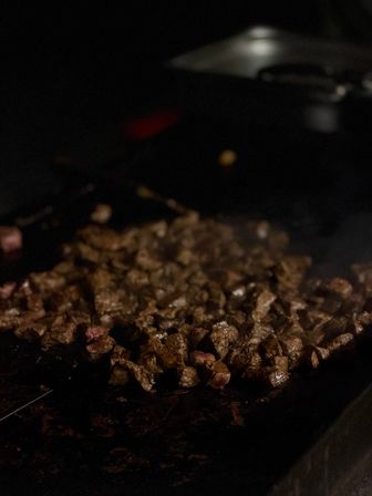 Close-up of sizzling diced steak bites browning on a hot flat-top griddle in a dimly lit restaurant kitchen.