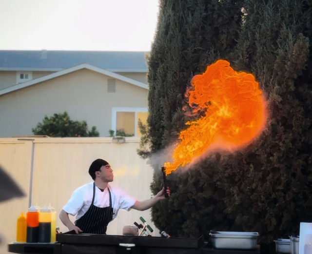 Cook in apron using a handheld blowtorch at an outdoor grill, sending a dramatic orange flame toward tall trees in a backyard setting with condiment bottles and prep pans on the cart.
