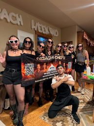 Group of women in black outfits and heart-shaped sunglasses holding a hibachi chef banner with a kneeling chef in a cozy wood-accented kitchen, 'bachelorette weekend' balloons overhead