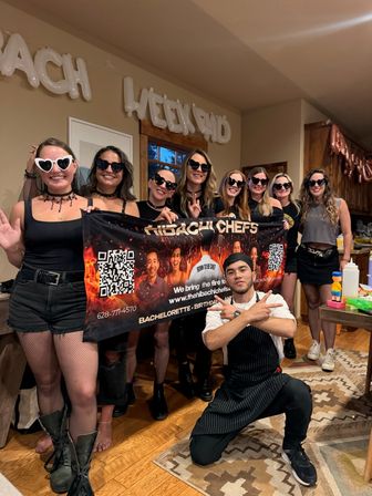 Group of women in black outfits and heart-shaped sunglasses holding a hibachi chef banner with a kneeling chef in a cozy wood-accented kitchen, 'bachelorette weekend' balloons overhead