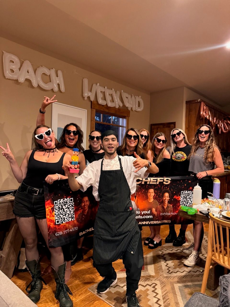 Smiling private chef in apron holds a small toy while posing with a laughing group of women in black sunglasses at a bachelorette weekend party in a cozy wood‑accented kitchen with balloon letters and a catering banner.