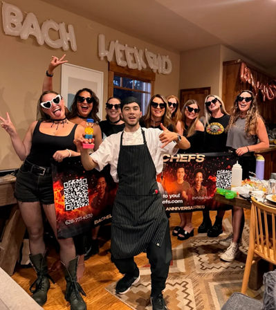 Bachelorette weekend party in a cozy home kitchen — group of women in black and heart-shaped sunglasses posing with a chef in an apron holding a novelty toy under 'BACH WEEKEND' balloons.
