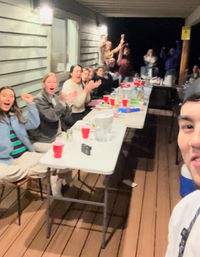 Friends cheering on a covered wooden porch at night, gathered around a long folding table with red cups, pitchers and snacks in a selfie-style group photo.